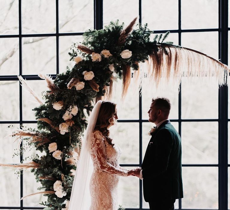 Winter wedding with cosy lighting and white rose wedding moongate with pampas grass decorating the altar with the bride in a lace wedding dress standing with the groom 