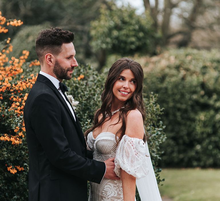 The groom in a black tuxedo gazes at the bride in a fitted boho lace gown with detachable sleeves posing for their couple portraits 