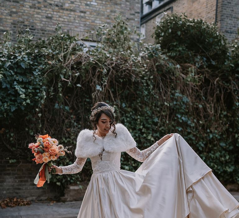 Bride in taffeta material princess wedding dress with long lace sleeves  and cropped faux fur bridal cover up with gold, pearl and emerald bridal crown holding bouquet with light pink poppies, orange garden roses, eucalyptus, Christmas red berry garlands, dried flowers and sage green and orange ribbon