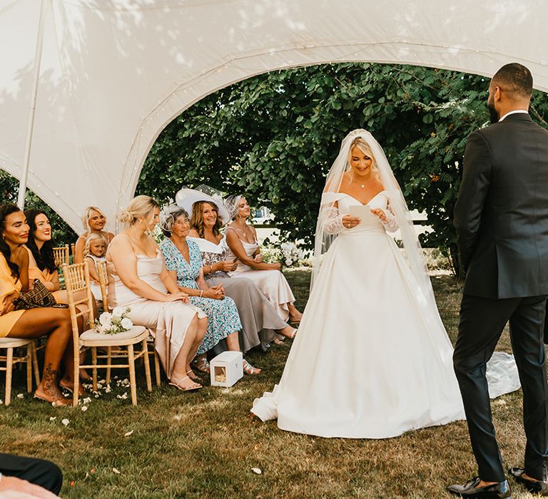 Blonde bride in princess wedding dress and drop veil reads her vows during tent wedding ceremony