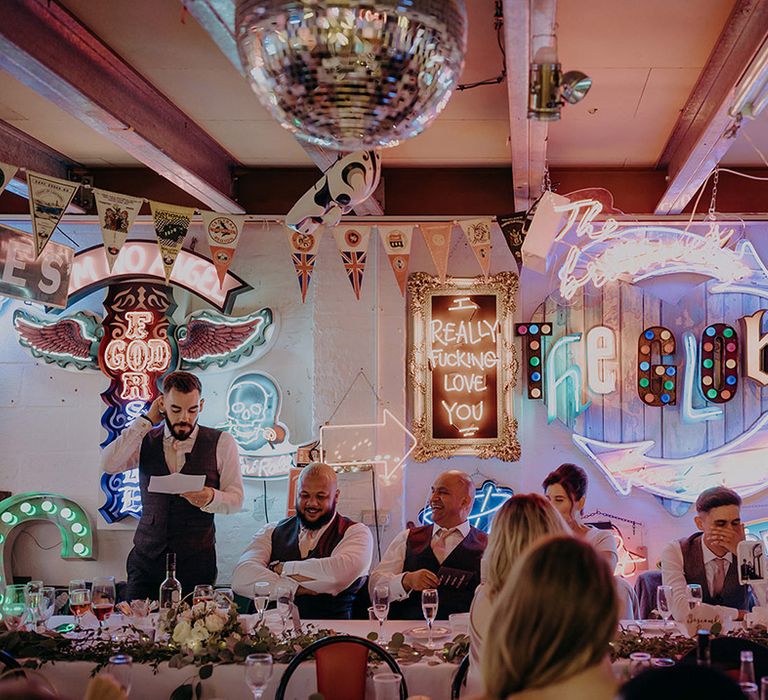 Groomsman making speech wearing dark grey waistcoat and yellow tie at the top table of London wedding venue reception room with neon signs, disco ball decor, bunting and LED signs 