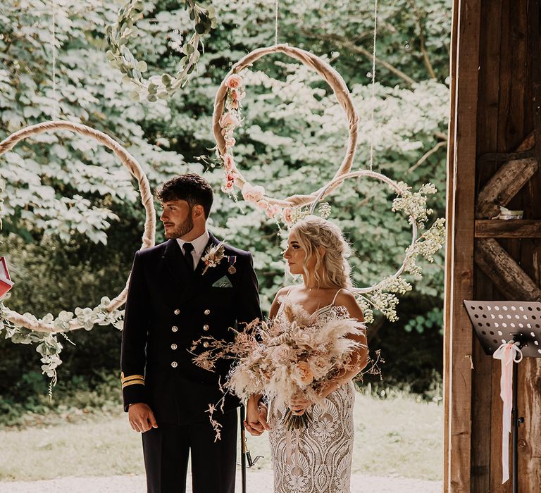 Hanging hula hoop decor for the altar decoration with foliage with the groom in a military uniform with the bride in a boho gown with a pampas grass bouquet matching the groom's buttonhole