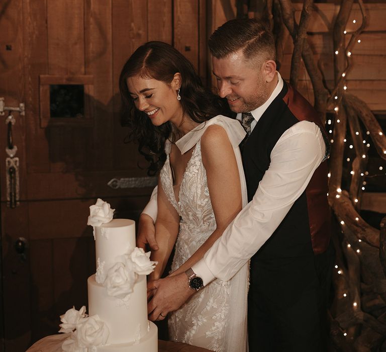 The bride and groom at their rustic wedding cut the three tier white wedding cake with white rose design 