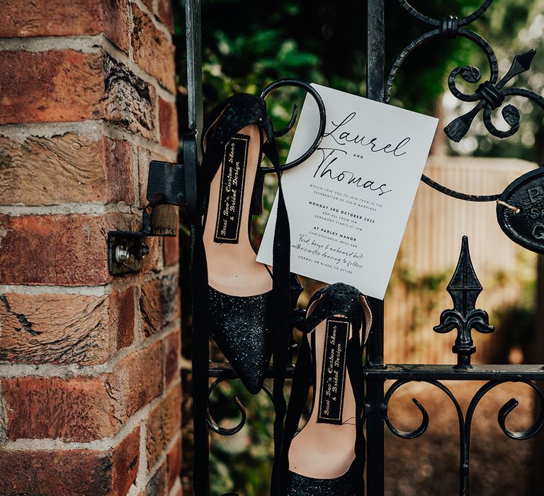 Black glitter pointed wedding shoes with ribbon to tie the shoe and black and white wedding stationery on a metal gate 