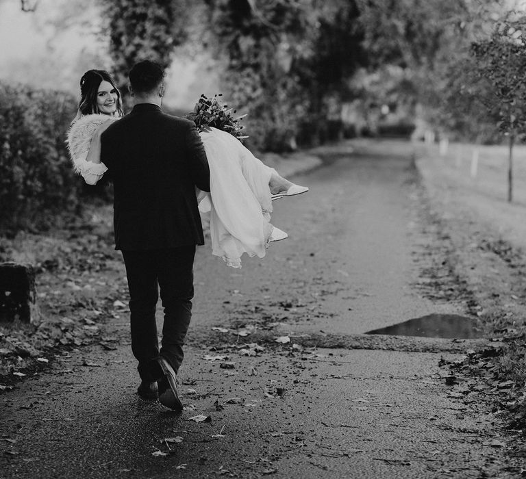 The groom walks along carrying the bride as they walk around the muddy wedding venue on a rainy day 