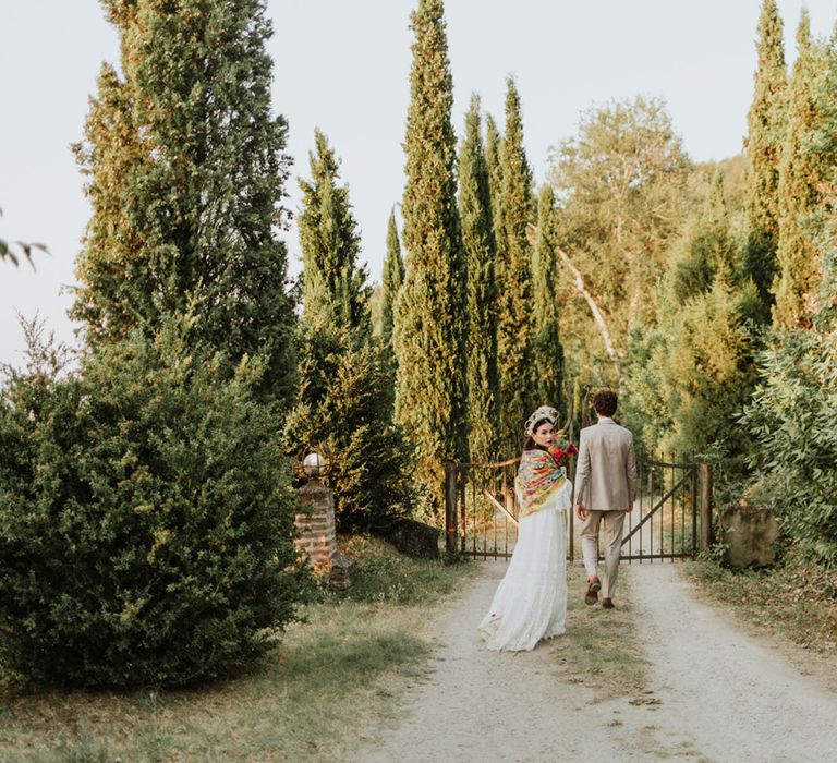 Bride and groom walk through greenery at italian destination wedding wearing white lace wedding dress, white floral structured bridal crown and greige suit