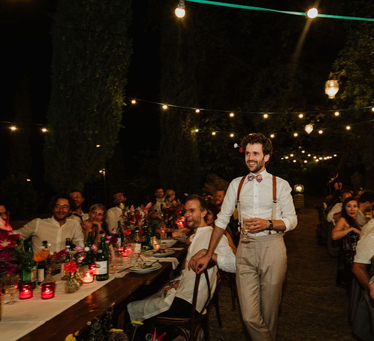 Groom in burnt orange braces holds glass of champagne at wedding reception in Italy