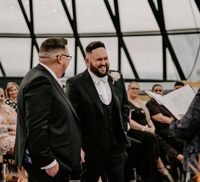 Grooms in matching classic black three piece tuxedos with white rose boutonnieres at the alter with wedding celebrant at The Gherkin London 