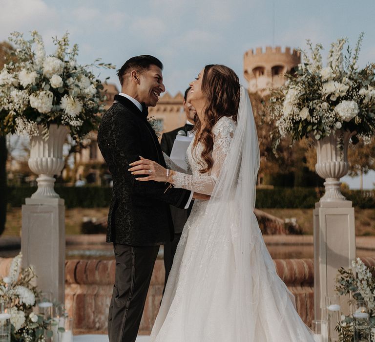 Bride in lace long sleeve Pronovias wedding dress and church-length veil embracing groom in black tuxedo with black bow tie and white carnation and dried flower boutonniere at alter of castle wedding venue Barcelona 