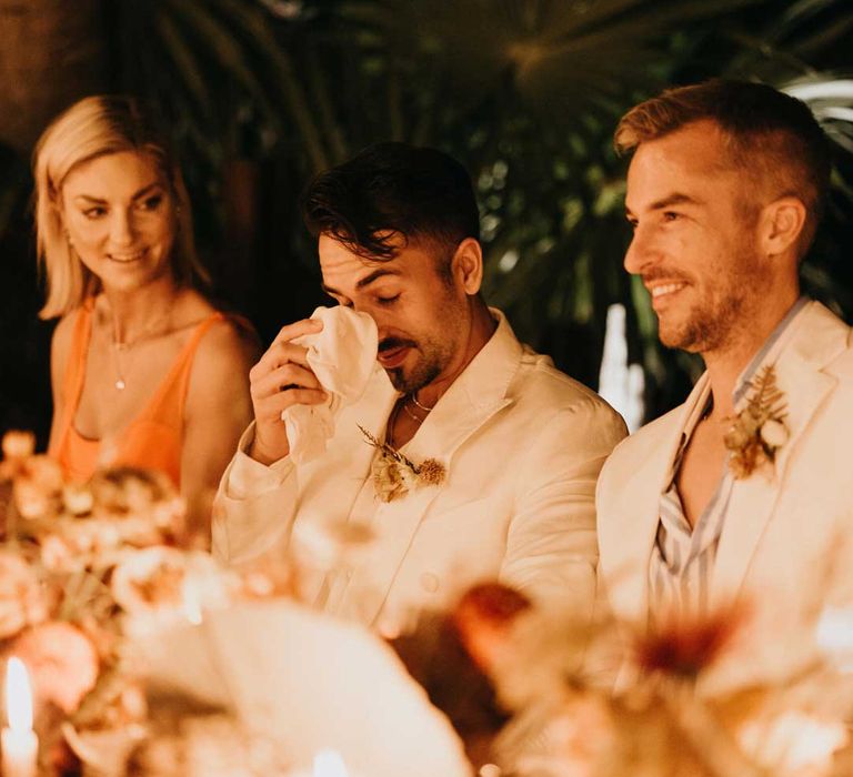 Groom in mens white wedding suit wiping a tear away during wedding speeches at destination wedding 