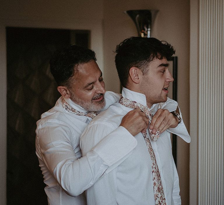 Groom in white shirt and floral patterned tie helping his son put on his patterned tie ready for the wedding day 