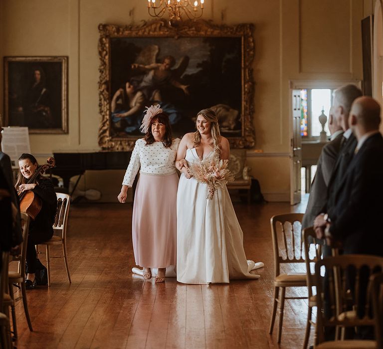 Mother of the bride in a white and pink wedding outfit walks the bride in a strapless Suzanne Neville wedding dress down the aisle at Doddington Hall and Gardens