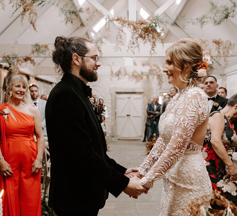 Bride in boho Catherine Deane wedding dress holding hands with the groom in a black velvet suit at the altar 