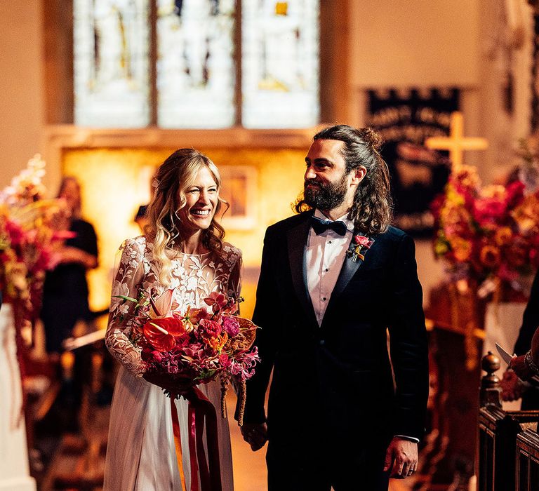 Groom with long brown hair with beard in black tie suit and bride in lace wedding dress holding colourful bright bouquet 