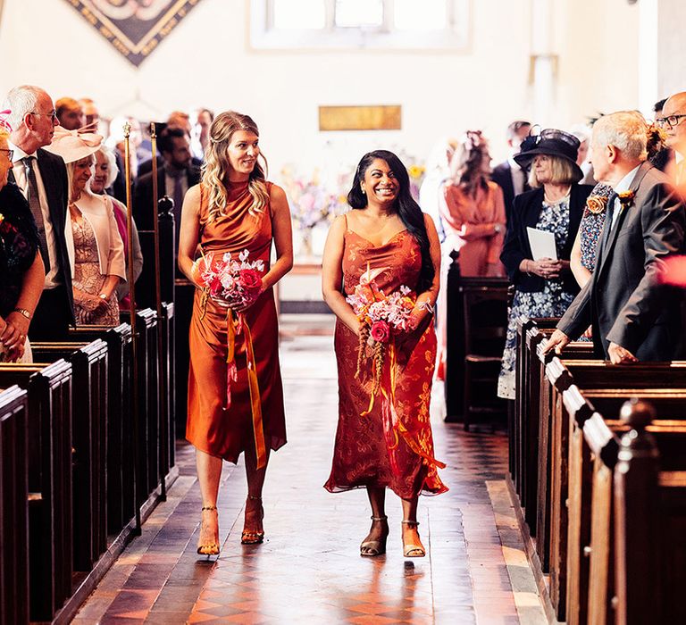 Bridesmaids in mismatched satin orange bridesmaid dresses walking down the aisle together 