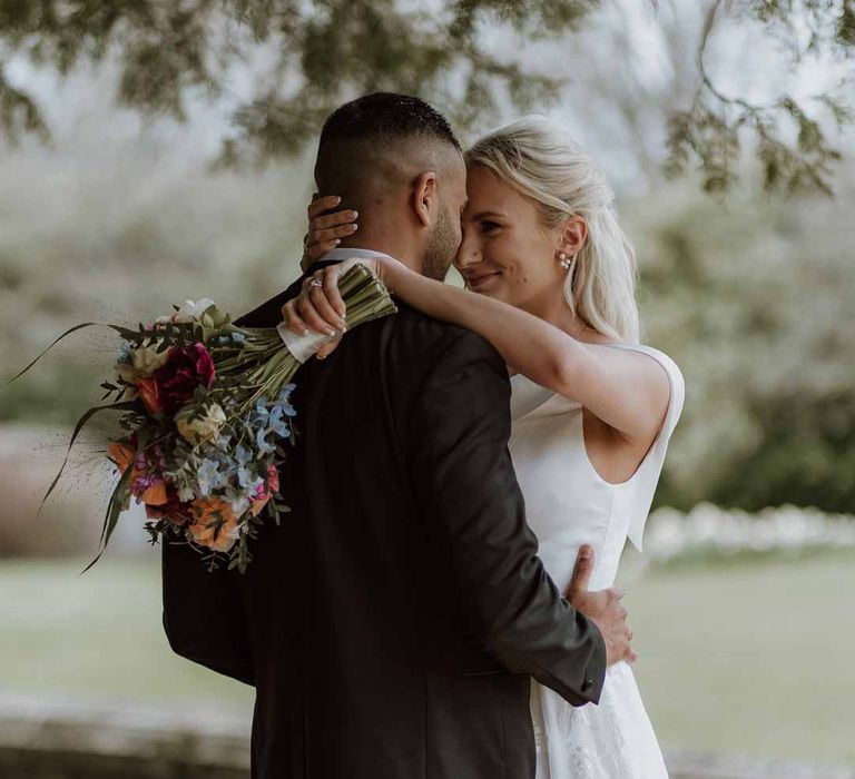 Bride in off the shoulder lace wedding dress holding colourful spring bridal bouquet embracing groom in classic three piece grooms tuxedo at Frensham Hall wedding venue