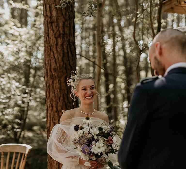 Bride holding purple and white wildflower wedding bouquet with floral lace off the shoulder wedding dress with detachable sleeves smiling up at the groom 