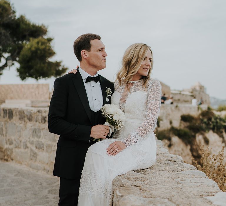 Ibiza destination wedding with bride in a lace wedding dress sitting on a wall holding an all white wedding bouquet with her groom in a tuxedo stood beside her