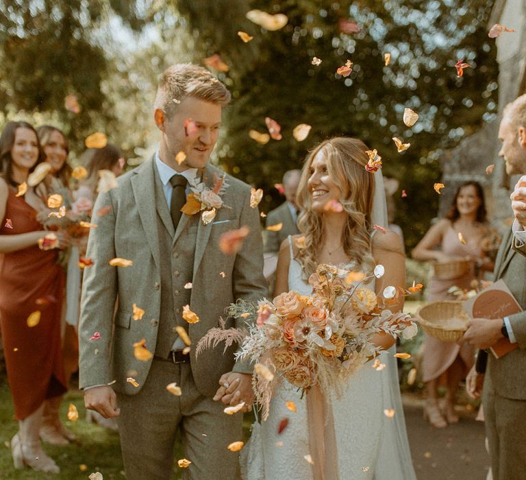 Groom in three piece wedding suit walking with the bride in classic fitted lace wedding dress celebrating their wedding with petal confetti 