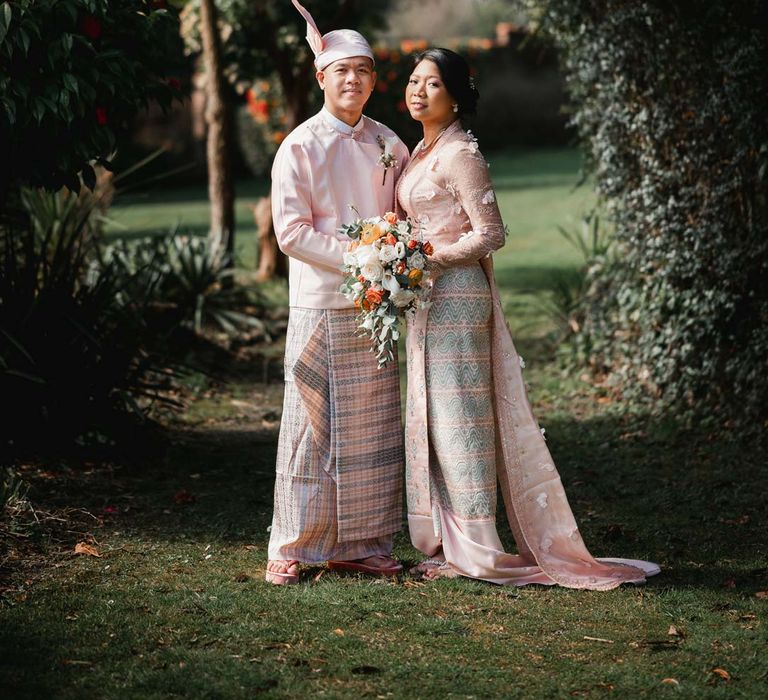 Bride in floral 3D applique pink and rose gold htamane standing on the grounds of Welsh wedding venue Bron Eifion with groom in baby pink htaingmathein 