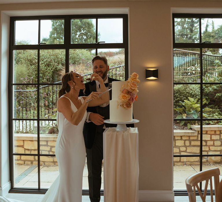 Bride and groom cut the two tier white frosted wedding cake decorated with pink and orange roses 