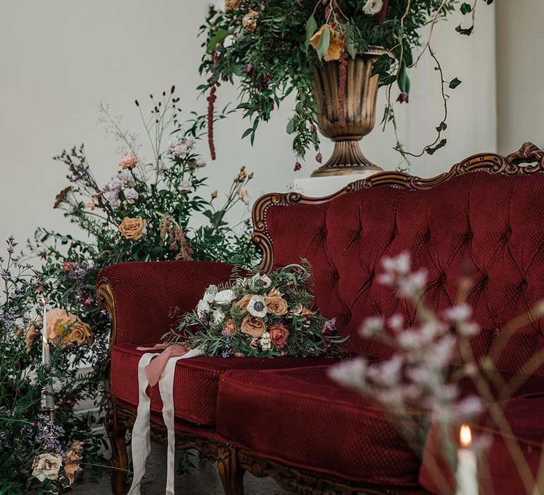 Deep maroon velvet loveseat surrounded by tapered candles and suspended wildflower and foliage arrangements in large gold vases at Buxted Park