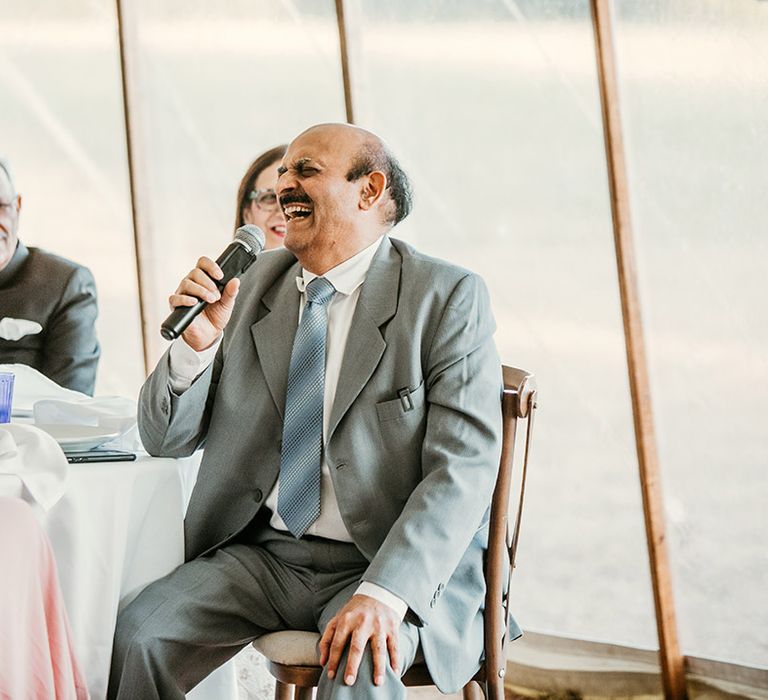 Father of the bride sitting in a blue suit with a microphone reading out his wedding speech 