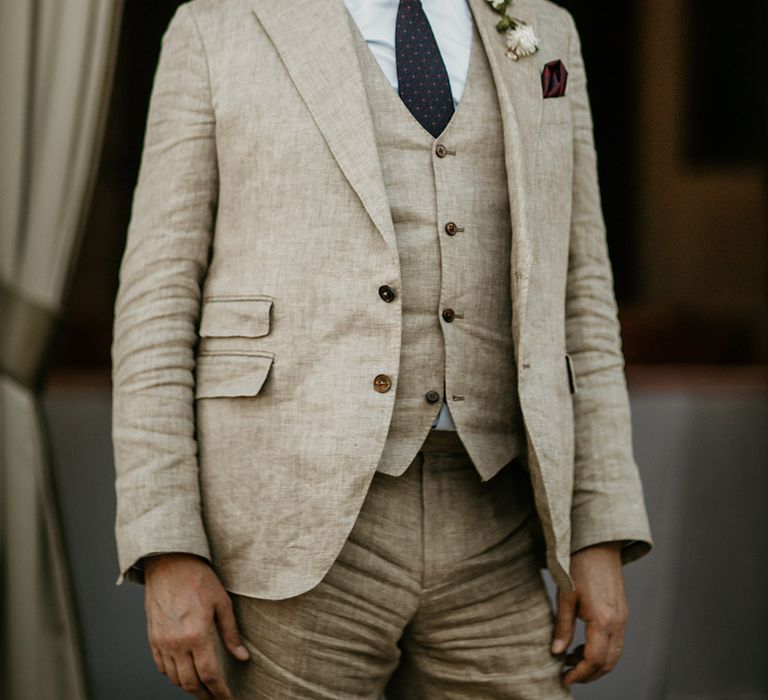 Groom in three piece cream wedding suit with dark blue wedding tie wearing glasses 