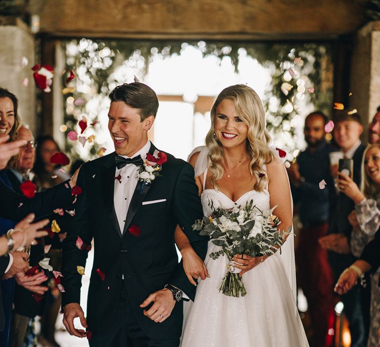 Groom in black tuxedo with bride in sparkly wedding dress walking back down the aisle for rose petal confetti moment 