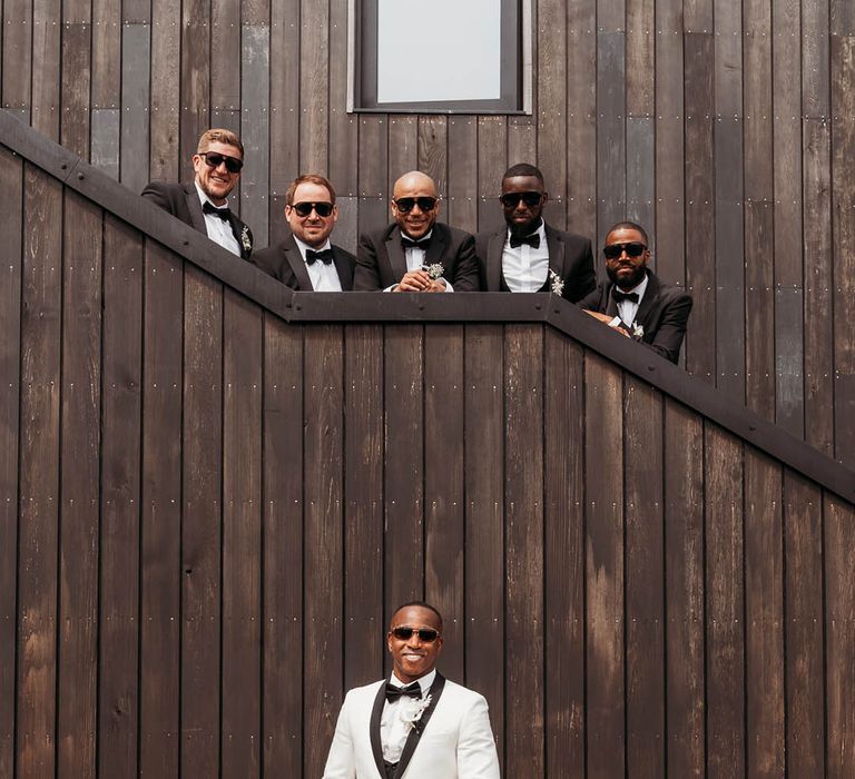 Groom in a white tuxedo jacket, sunglasses and black bow tie stands under groomsmen in black groomsmen suits and sunglasses