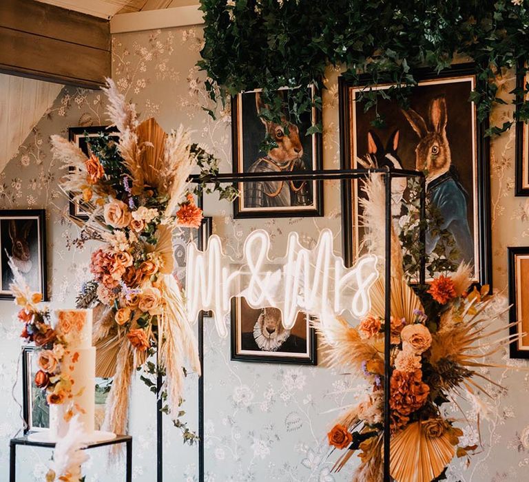 Neon Mr & Mrs white wedding sign on black metal arch decorated with orange autumnal flowers at the ceremony 