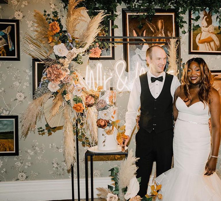 Bride in mermaid wedding dress posing with groom next to their three tier wedding cake covered in autumnal flowers and neon sign 