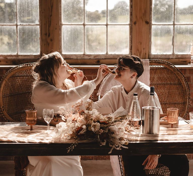 Brides feed each other pizza while sitting at their sweetheart table with peacock chairs 