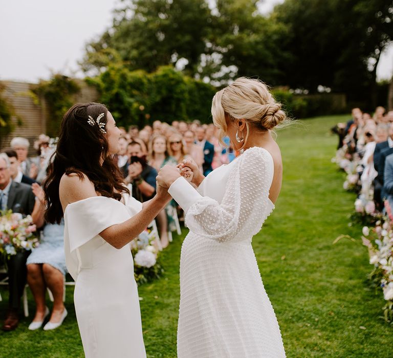 Two brides holding hands together at their same sex outdoor wedding ceremony 