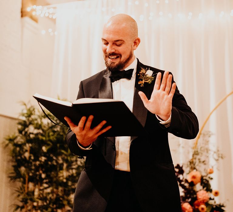 Best man in black tuxedo reading out wedding speech at rustic barn wedding 