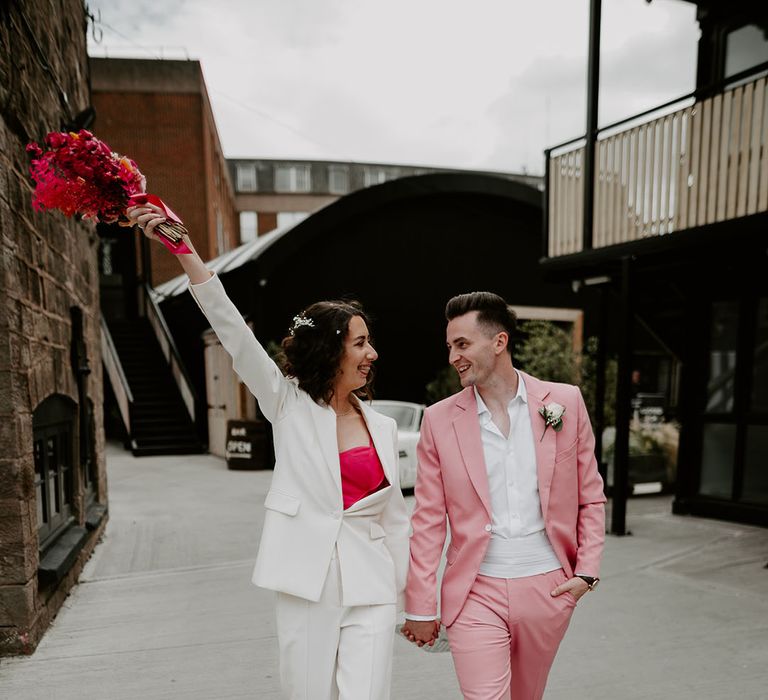 The bride wearing a white bridal suit with heart shaped pink top walks with the groom in a pink suit 