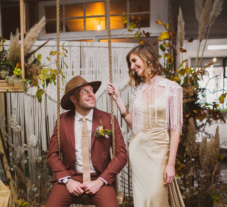 Bride in gold dress and groom sitting on a swing wearing a burgundy suit and hat