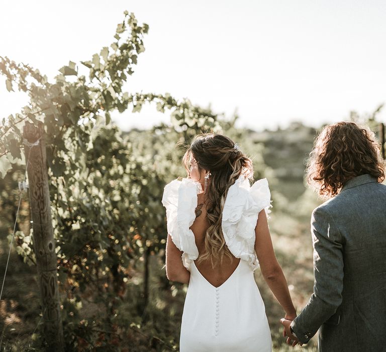 Bride in low back wedding dress with button detail and ruffle sleeves holding her grooms hand in a blue suit in a vineyard in Italy 