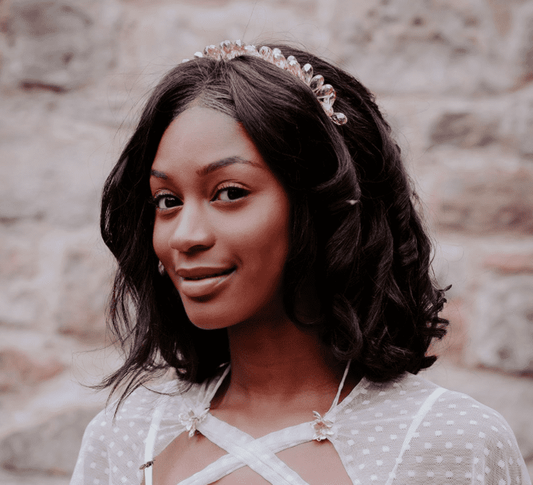 A Black bride looks coyly towards the camera. She has a dress with a cape on and short loosely waved hair.