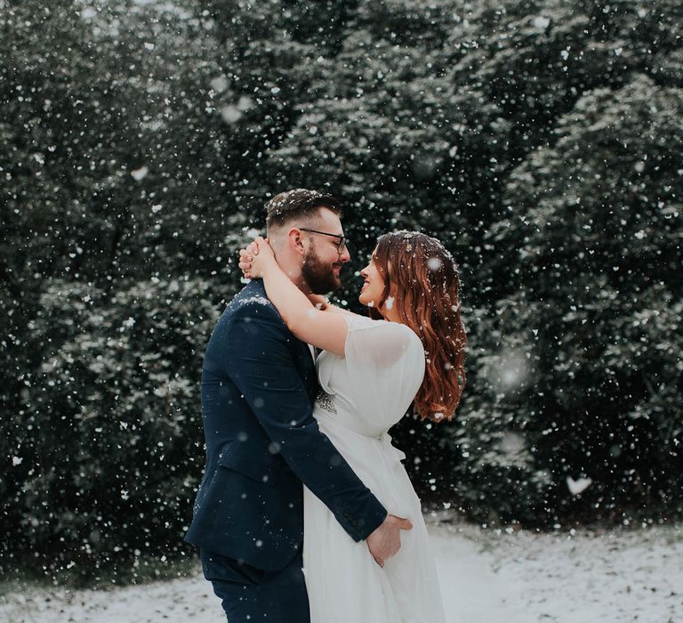 Groom in navy suit swings bride in white dress at snowy wedding