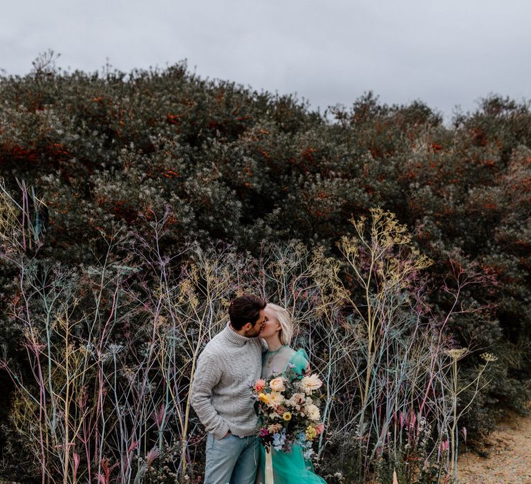 Groom in a grey turtle neck jumper kissing his bride in a green sheer wedding dress at their intimate beach elopement with colourful flowers and picnic area 