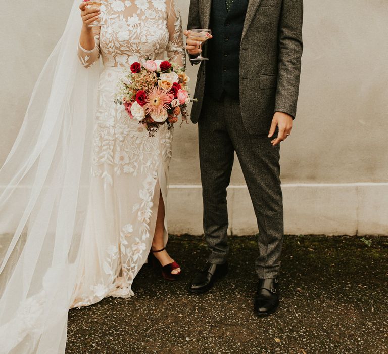 Bride in a lace Hermione de Paula wedding dress with front split and red velvet shoes holding a red autumn wedding bouquet standing next to her groom in a grey wool suit 