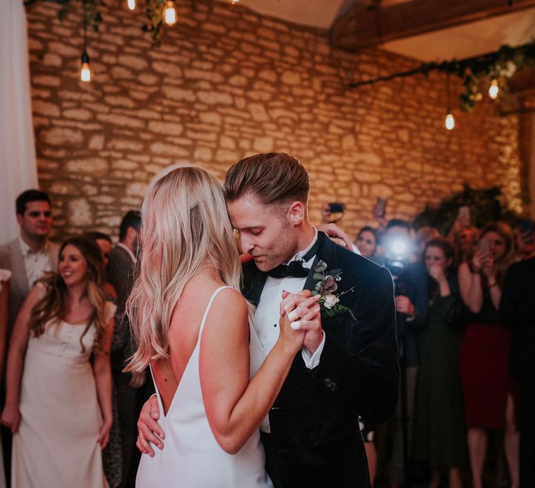 Bride in white silk v back dress dances with groom in black velvet Hugo Boss suit in stone barn with foliage decor at Caswell House wedding