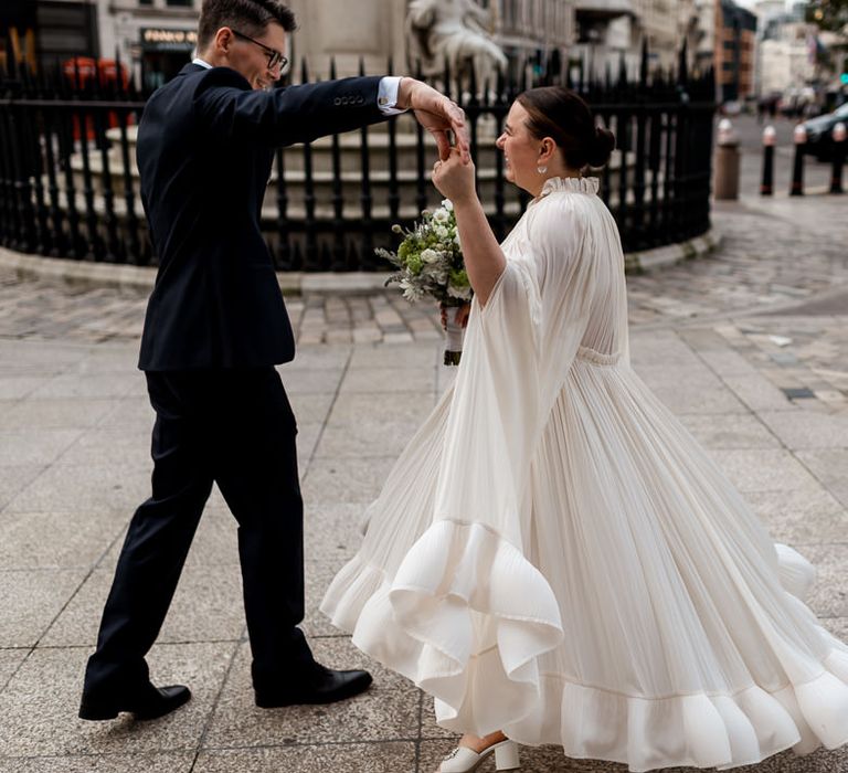 Groom in a navy suit twirling his bride in a Lanvin wedding dress and bridal cape with long sleeves and ruffle hem