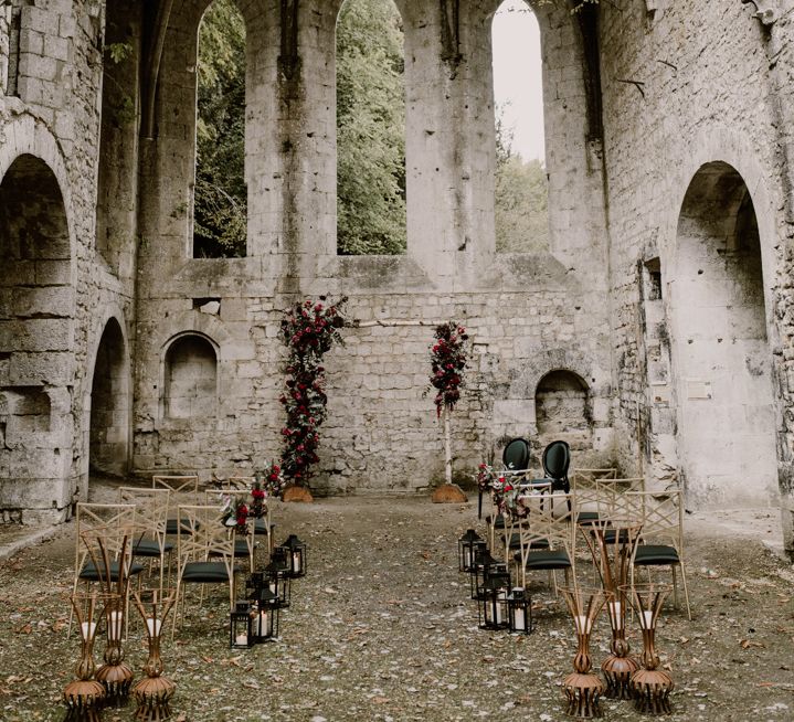 Gothic style wedding ceremony in an open air medieval abbey with red flower arch and aisle chairs 