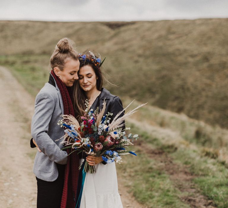 The brides cuddling in the Peak District, holding a bright bouquet of red and blue flowers