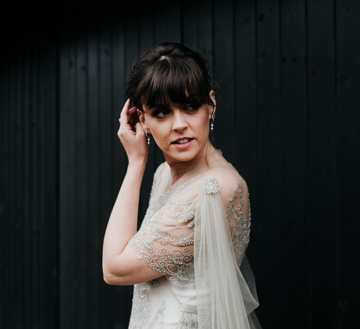 A bride with a lonf fringe brushes her hair out of her eyes. She wears a sheer cape.