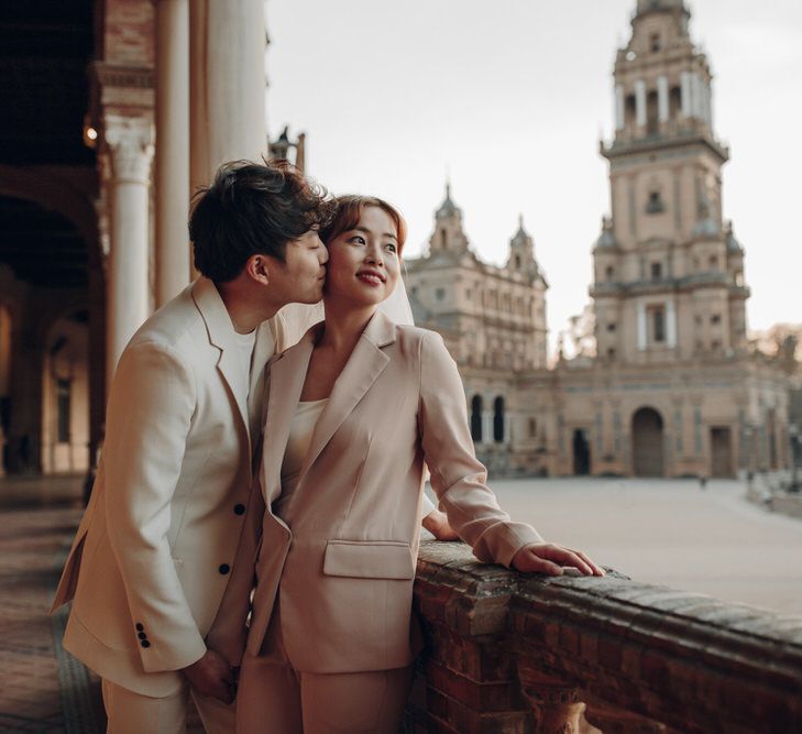 Asian woman wearing veil and pink suit being kissed on cheek by partner wearing cream suit in Seville