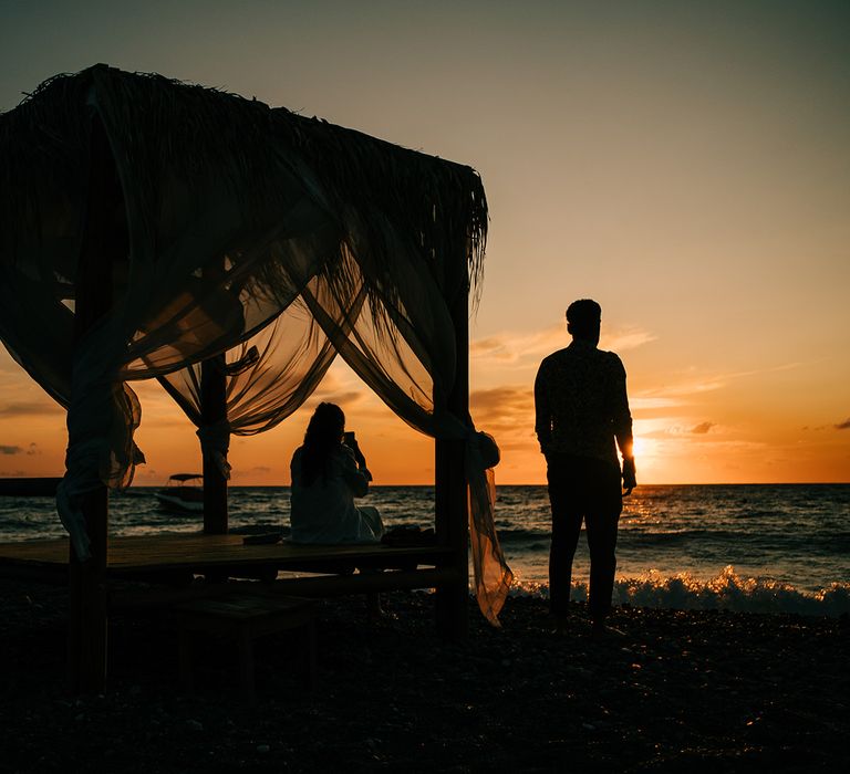 Couple sit on the beach during sunset in Greece for honeymoon photoshoot