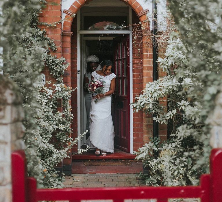 Bride in a fitted lace wedding dress holding a red and green bouquet walking out of the front door